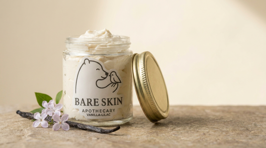 Vanilla-Lilac Body Butter by Bare Skin Apothecary - whipped ivory body butter in a clear glass jar with gold lid and white die-cut label, vanilla bean and lilac blossoms